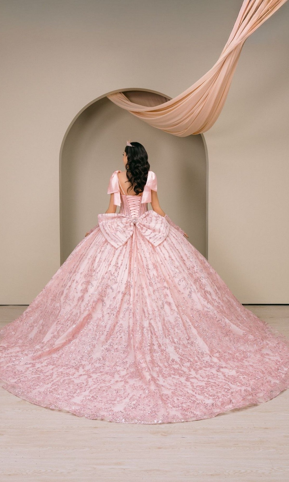 A woman in a pink Quinceanera Dress 8530JQ By Cinderella Couture, featuring a beaded corset bodice, stands facing away in front of an arched doorway with draped fabric overhead, evoking timeless elegance.